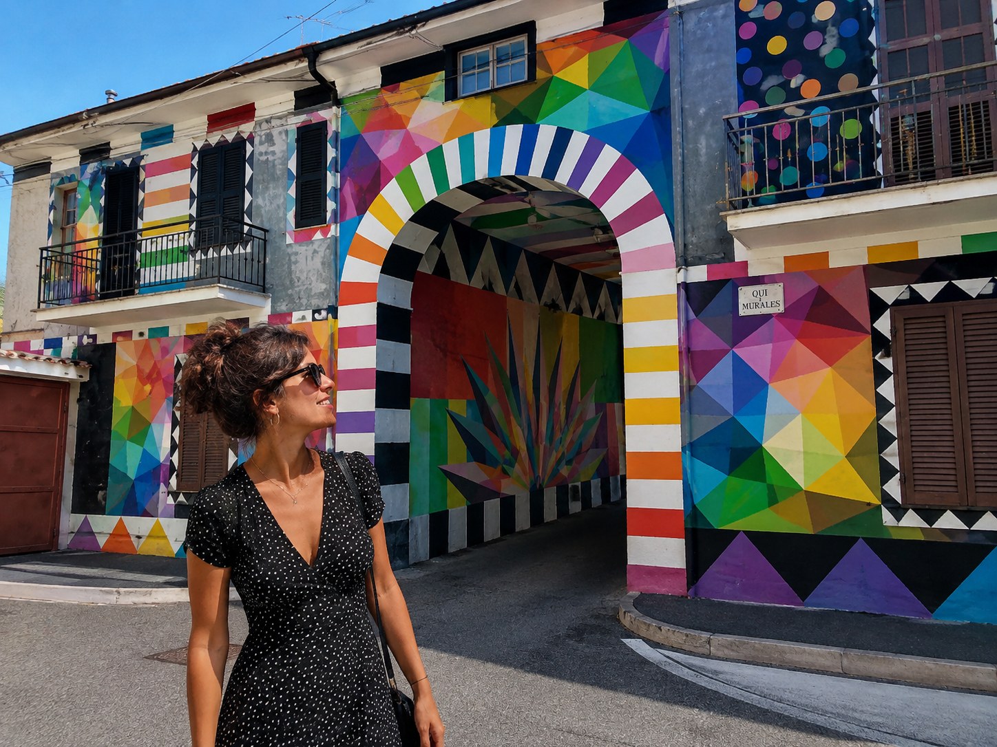 Passante devant une façade d'Aielli entièrement recouverte d'une murale géométrique aux couleurs vives, style Okuda San Miguel, avec un passage voûté peint à rayures multicolores.