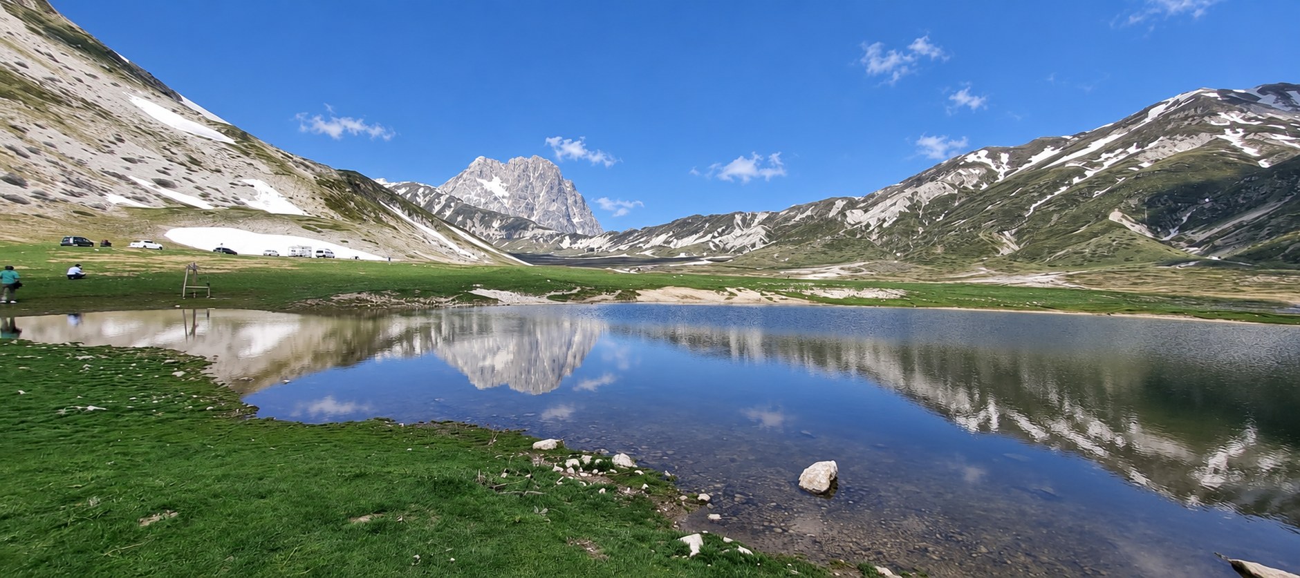 Vue panoramique du lac Pietranzoni, parfaitement calme, reflétant le Corno Grande, le ciel bleu et les montagnes environnantes, avec des camping-cars garés sur le plateau de Campo Imperatore.