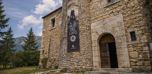 Illustration de la façade en pierre du Museo del Lupo à Rocca di Cambio, avec sa tour et sa bannière consacrée au loup des Apennins.