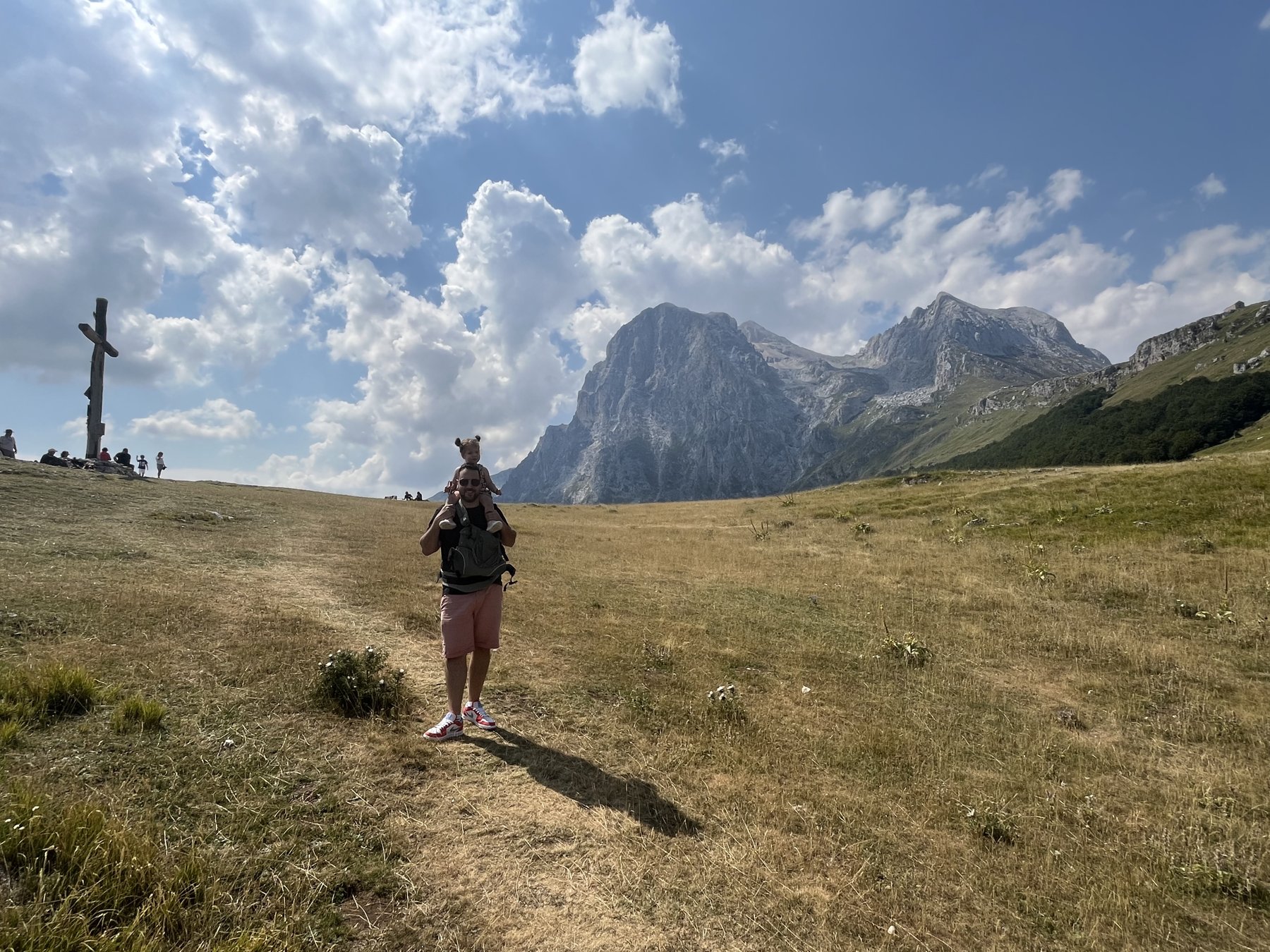 Un père porte son enfant sur les épaules dans la prairie de Prati di Tivo, face aux sommets du Gran Sasso, avec la croix de Cima Alta visible à gauche.