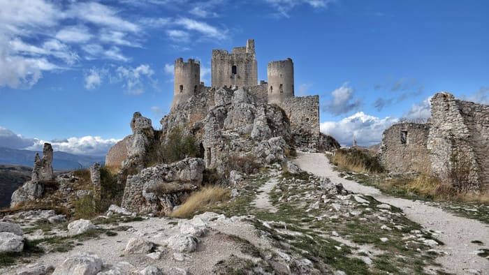 Le sentier menant à Rocca Calascio en pleine journée, bordé des vestiges du village abandonné, avec la forteresse en arrière-plan sous un ciel bleu.